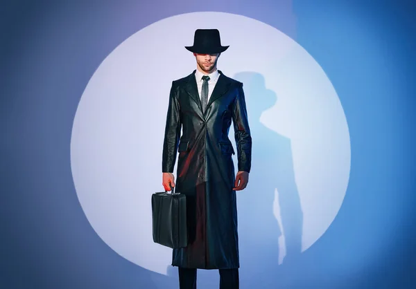 Full length portrait of handsome man in black coat and hat holding briefcase posing in the spotlight on studio background — Fotografia de Stock