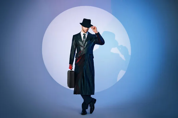 Full length portrait of handsome man in black coat and hat holding briefcase posing in the spotlight on studio background —  Fotos de Stock