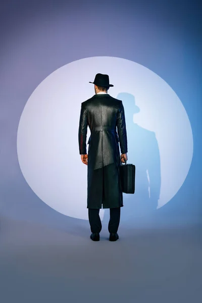 Back view of man silhouette in black coat and hat holding briefcase in the spotlight on studio background — Stock Photo, Image