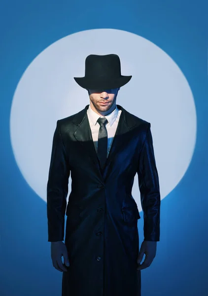 Portrait of handsome man in black coat hiding face behind his hat in the spotlight on studio background — 스톡 사진