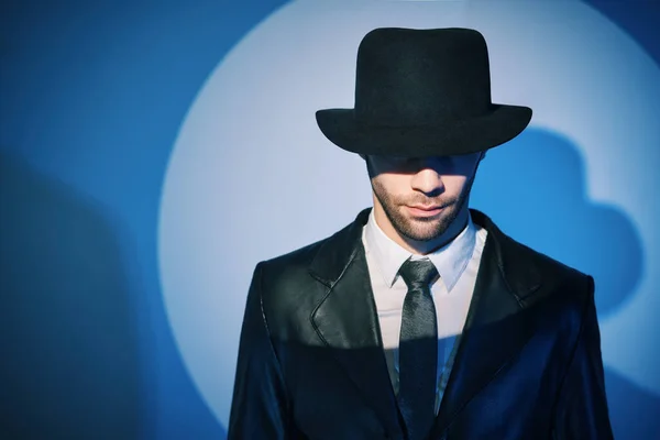 Portrait of handsome man in black coat hiding face behind his hat in the spotlight on studio background — Fotografia de Stock