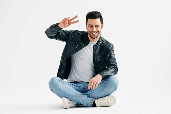Positive cheerful young man doing victory sign sitting with legs crossed on floor isolated over white background — стоковое фото