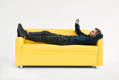 Sarı kanepede uzanmış rahat bir adam beyaz arka planda cep telefonuyla görüntülü konuşma yapıyor. Sosyal medya kavramı