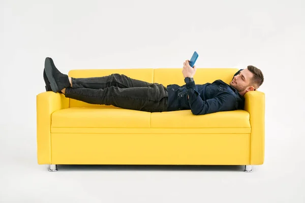 Relaxált Férfi Fekvő Sárga Kanapén Okostelefon Segítségével Fehér Háttér — Stock Fotó