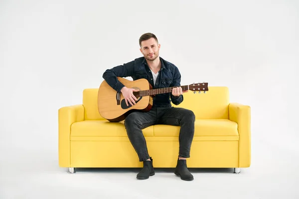 Przystojny Młody Człowiek Gra Gitarze Akustycznej Siedząc Żółtej Kanapie Białym — Zdjęcie stockowe