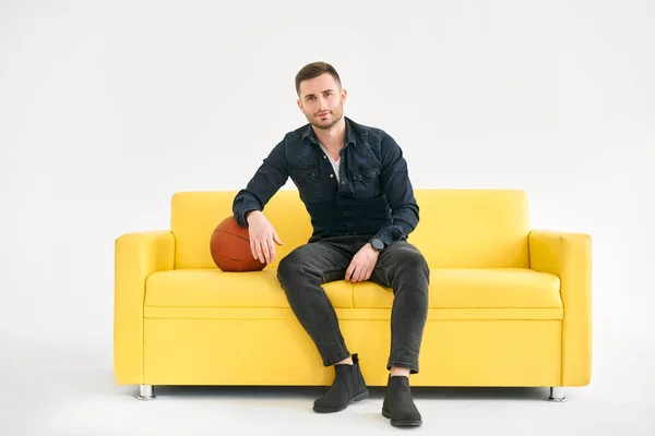 年轻的时髦男子坐在黄色沙发上的篮球白色背景 体育概念 — 图库照片