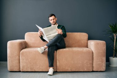 Confident young man reading newspaper and latest news while sitting on sofa in the morning in modern interior