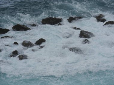 Beyaz köpüklü dalgalar sahile yakın kayalara çarpıyor. 