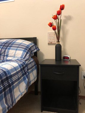 Çiçek vazosu ve çalar saati olan komodinin yanında.