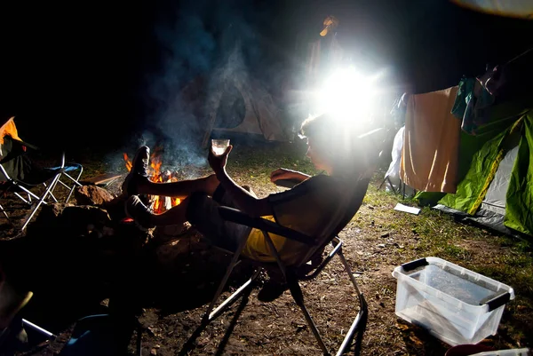 男が火事で椅子に座っている キャンペーンで夜の休息 火とテントを背景にアルコールのガラスを持つ男のシルエット 芸術的効果としての劇的な光との対比 — ストック写真