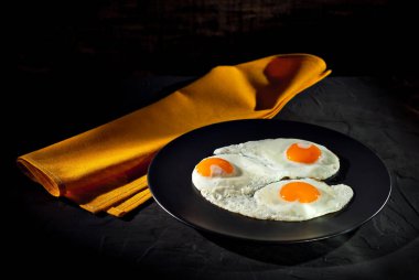 Siyah tabakta çırpılmış yumurta. Eski bir beton masada sahanda yumurta. Tabağın yanındaki sarı masa örtüsü..