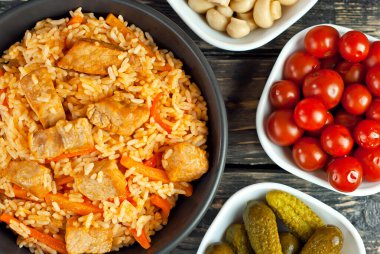 Siyah kasede et ve havuçlu pilav. Siyah arka planda marine edilmiş mantar, domates ve salatalık. Sehpada yemek. Eski ahşap bir tahtada kase.