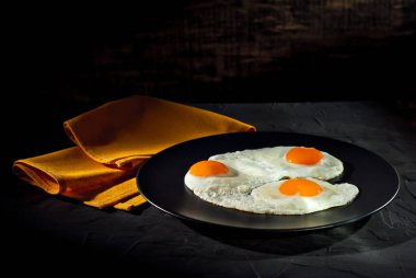 Siyah tabakta çırpılmış yumurta. Eski bir beton masada sahanda yumurta. Tabağın yanındaki sarı masa örtüsü..