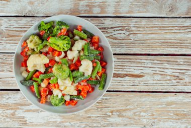 Beyaz bir kasede donmuş sebze karışımı. Eski püskü bir masada yemek hazırlığı. Işık panosu arka planında kesilmiş sebzeler. Yiyeceğin yanındaki metin için boşluk ve boşluk kopyala.