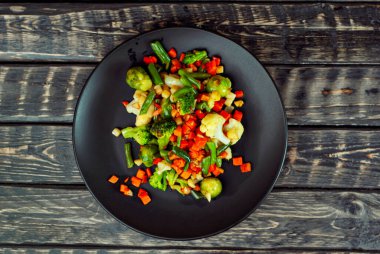 Sebze karışımı. Çeşitli sebze salatası. Kızarmış lahana, havuç, brokoli, siyah tabakta bezelye. Eski ahşap bir tahtanın arkasında yemek..