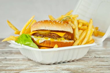 Eski püskü bir masada beyaz tabakta çizburger. Hamburger ve patates kızartması yakın mesafede. İşlenmiş mısırdan yapılmış ekolojik bir konteynırda yiyecek. Çevre dostu yemekler.