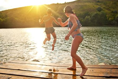 Çocuklar iskeleden suya atlıyor. Suyun kenarında kamp yapmak. Günbatımı arka planında aktif dinlenme. Yaz, tatil, seyahat ve tatil kavramı.
