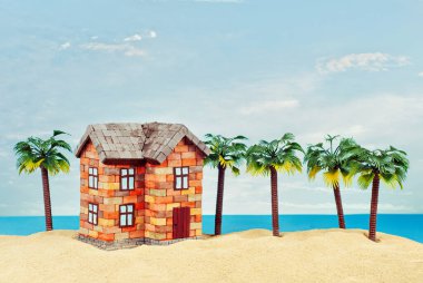 Kumda bir ev maketi. Deniz ve mavi gökyüzünün arka planında bir sahil evi. Ev ve ipotek konsepti. Palmiye ağaçları ve evin yakınındaki cennet adası..