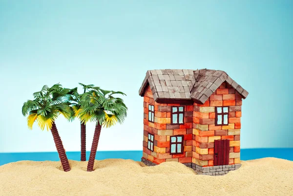 Kumda bir ev maketi. Deniz ve mavi gökyüzünün arka planında bir sahil evi. Ev ve ipotek konsepti. Palmiye ağaçları ve evin yakınındaki cennet adası..