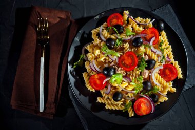 Domatesli salata, biber, ton balığı ve makarna. Siyah tabakta, siyah arka planda taze salata. Sarı çatal tabağın yanında..