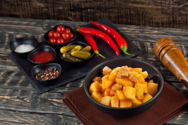 Siyah kasede etli haşlanmış patates. Siyah ahşap bir masada baharatlar ve baharatlar. Eski püskü bir arka planda ketçap, tuz ve biber..