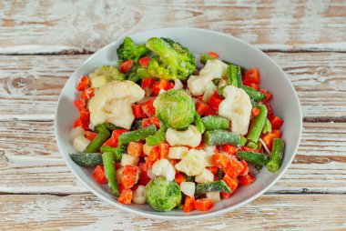 Beyaz bir kasede donmuş sebze karışımı. Eski püskü bir masada yemek hazırlığı. Dilimlenmiş sebzeler arka planda.