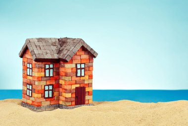 Kumda bir ev maketi. Deniz ve mavi gökyüzünün arka planında bir sahil evi. Ev ve ipotek konsepti. Evin yakınındaki cennet adası..