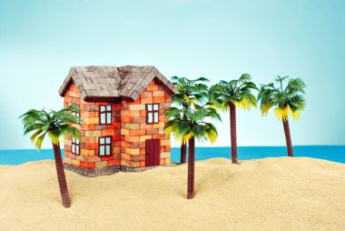 Kumda bir ev maketi. Deniz ve mavi gökyüzünün arka planında bir sahil evi. Ev ve ipotek konsepti. Palmiye ağaçları ve evin yakınındaki cennet adası..