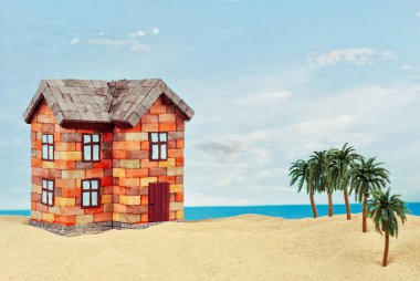 Kumda bir ev maketi. Deniz ve mavi gökyüzünün arka planında bir sahil evi. Ev ve ipotek konsepti. Palmiye ağaçları ve evin yakınındaki cennet adası..