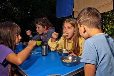 Çocuklar mavi bir kamp masasında yemek yiyorlar. Açık hava eğlencesi. Yeşillik arka planında çocuk pikniği.