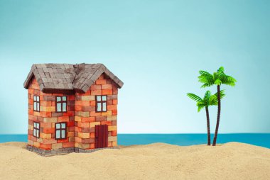 Kumda bir ev maketi. Deniz ve mavi gökyüzünün arka planında bir sahil evi. Ev ve ipotek konsepti. Palmiye ağaçları ve evin yakınındaki cennet adası..