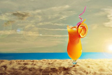 Plajda portakal kokteyli. Buzlu alkollü içecek, kumda portakal rengi. Arka planda gün batımı, deniz ve gökyüzü. Cama yakın metin için boşluk ve boş alanı kopyala.