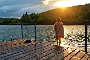 Havluya sarılmış bir kız rıhtımın kenarında duruyor. Eylemler suyun yakınındaki doğada dinleniyor. Arkaplanda gün batımı.