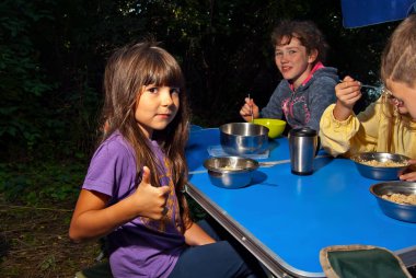 Çocuklar mavi bir kamp masasında yemek yiyorlar. Açık hava eğlencesi. Yeşillik arka planında çocuk pikniği.