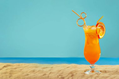 Plajda portakal kokteyli. Buzlu alkollü içecek, kumda portakal rengi. Arka planda gün batımı, deniz ve gökyüzü. Cama yakın metin için boşluk ve boş alanı kopyala.