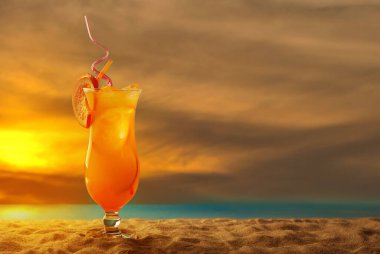 Plajda portakal kokteyli. Buzlu alkollü içecek, kumda portakal rengi. Arka planda gün batımı, deniz ve gökyüzü. Cama yakın metin için boşluk ve boş alanı kopyala.