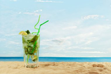 Kum üzerinde pipetle yeşil kokteyl. Plajda buzlu, limonlu ve naneli mojito. Arka planda gün batımı, deniz ve gökyüzü. Cama yakın metin için boşluk ve boş alanı kopyala.