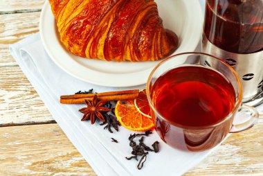 Beyaz tabakta kruvasan ve çay. Eski ahşap bir masada çay fincanı. Tam yaprak siyah çay, limon ve tarçın peçetede.. 