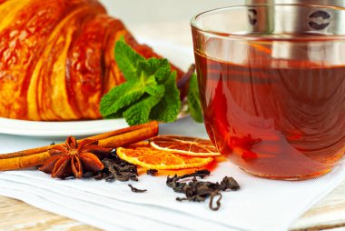 Beyaz tabakta kruvasan ve çay. Eski ahşap bir masada çay fincanı. Tam siyah çay, limon, nane yaprağı ve tarçın peçetede..