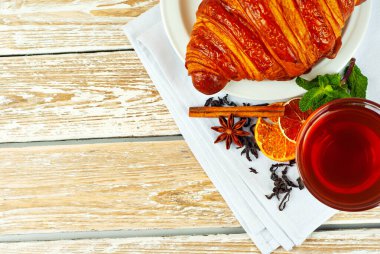 Beyaz tabakta kruvasan ve çay. Eski ahşap bir masada çay fincanı. Tam siyah çay, limon, nane yaprağı ve peçetede tarçın. Yemek yakınındaki metin için boşluk ve boş alanı kopyala.