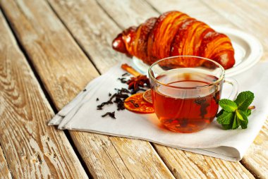 Beyaz tabakta kruvasan ve çay. Eski ahşap bir masada çay fincanı. Tam siyah çay, limon, nane yaprağı ve peçetede tarçın. Yemek yakınındaki metin için boşluk ve boş alanı kopyala.