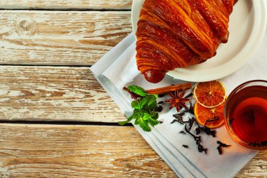 Beyaz tabakta kruvasan ve çay. Eski ahşap bir masada çay fincanı. Tam siyah çay, limon, nane yaprağı ve peçetede tarçın. Yemek yakınındaki metin için boşluk ve boş alanı kopyala.