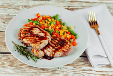 Kızarmış et parçaları. Biberiyeli biftek ve beyaz tabakta biber. Beyaz ahşap bir masada kırmızı soslu et yemeği. Bifteğin yanında mısır, havuç ve bezelye salatası..