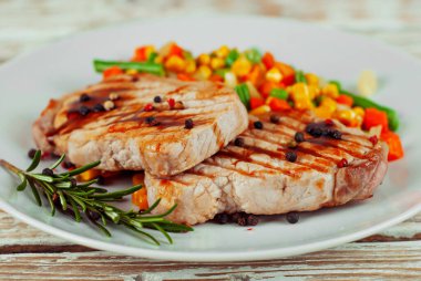 Kızarmış et parçaları. Biberiyeli biftek ve beyaz tabakta biber. Beyaz ahşap bir masada kırmızı soslu et yemeği. Bifteğin yanında mısır, havuç ve bezelye salatası..