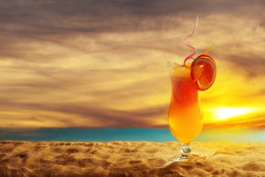 Plajda portakal kokteyli. Buzlu alkollü içecek, kumda portakal rengi. Arka planda gün batımı, deniz ve gökyüzü. Cama yakın metin için boşluk ve boş alanı kopyala.