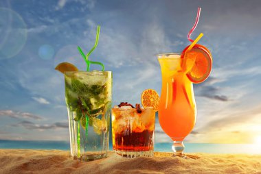 Plajda turuncu ve yeşil kokteyller. Buzlu, portakallı, naneli ve limonlu alkollü içecek. Arka planda gün batımı, deniz ve gökyüzü. Cama yakın metin için boşluk ve boş alanı kopyala.