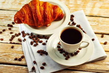 Beyaz seramik bardakta kahve. Kahvaltıda kruvasan ve kahve. Eski ahşap bir masada kahve. Kahve çekirdekleri fincanın yanına dağılmış..