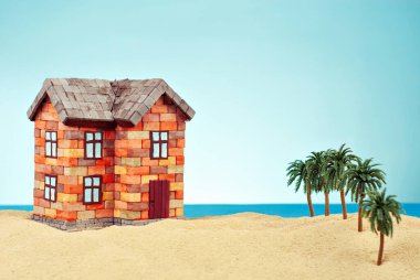 Kumda bir ev maketi. Deniz ve mavi gökyüzünün arka planında bir sahil evi. Ev ve ipotek konsepti. Palmiye ağaçları ve evin yakınındaki cennet adası..