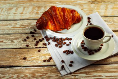 Beyaz seramik bardakta kahve. Kahvaltıda kruvasan ve kahve. Eski ahşap bir masada kahve. Kahve çekirdekleri fincanın yanına dağılmış..