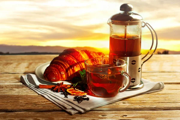 木製のテーブルの上にお茶とガラスティーポットのマグカップ 日没の背景にお茶とクロワッサン 白いナプキンに粗い紅茶 レモン シナモンとミントの葉 — ストック写真